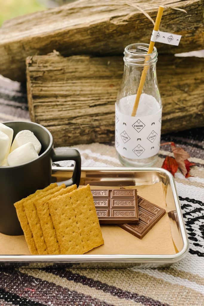 small portion of smores ingredients on mini metal cookie sheet next to glass milk bottle and wood logs