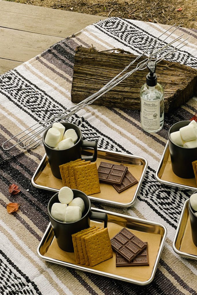 metal roasting sticks resting on picnic table next to plates of smores ingredients and hand sanitizer